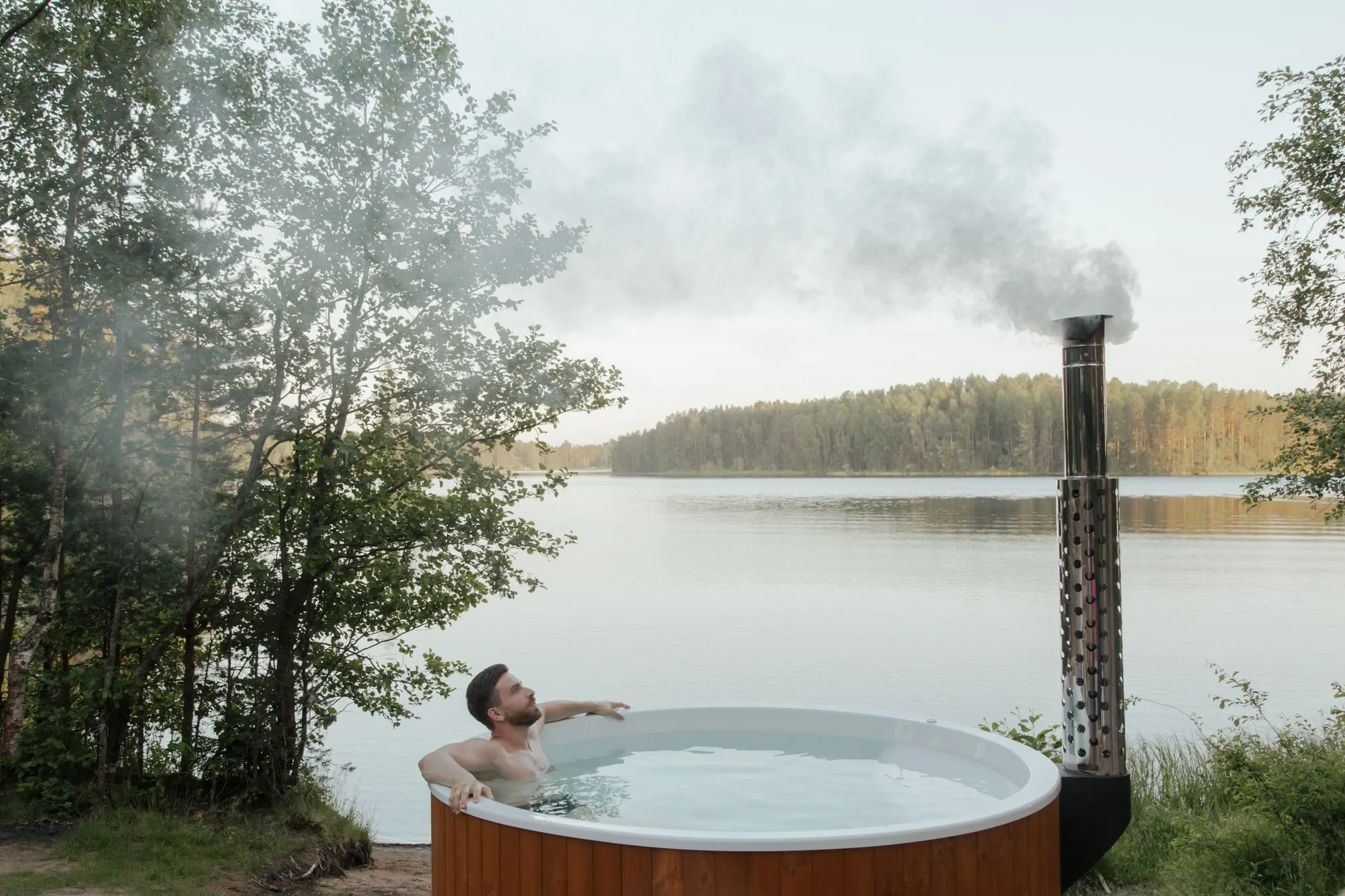 Homme dans un bain nordique en bois au bord d'un lac, la cheminée du poêle fume