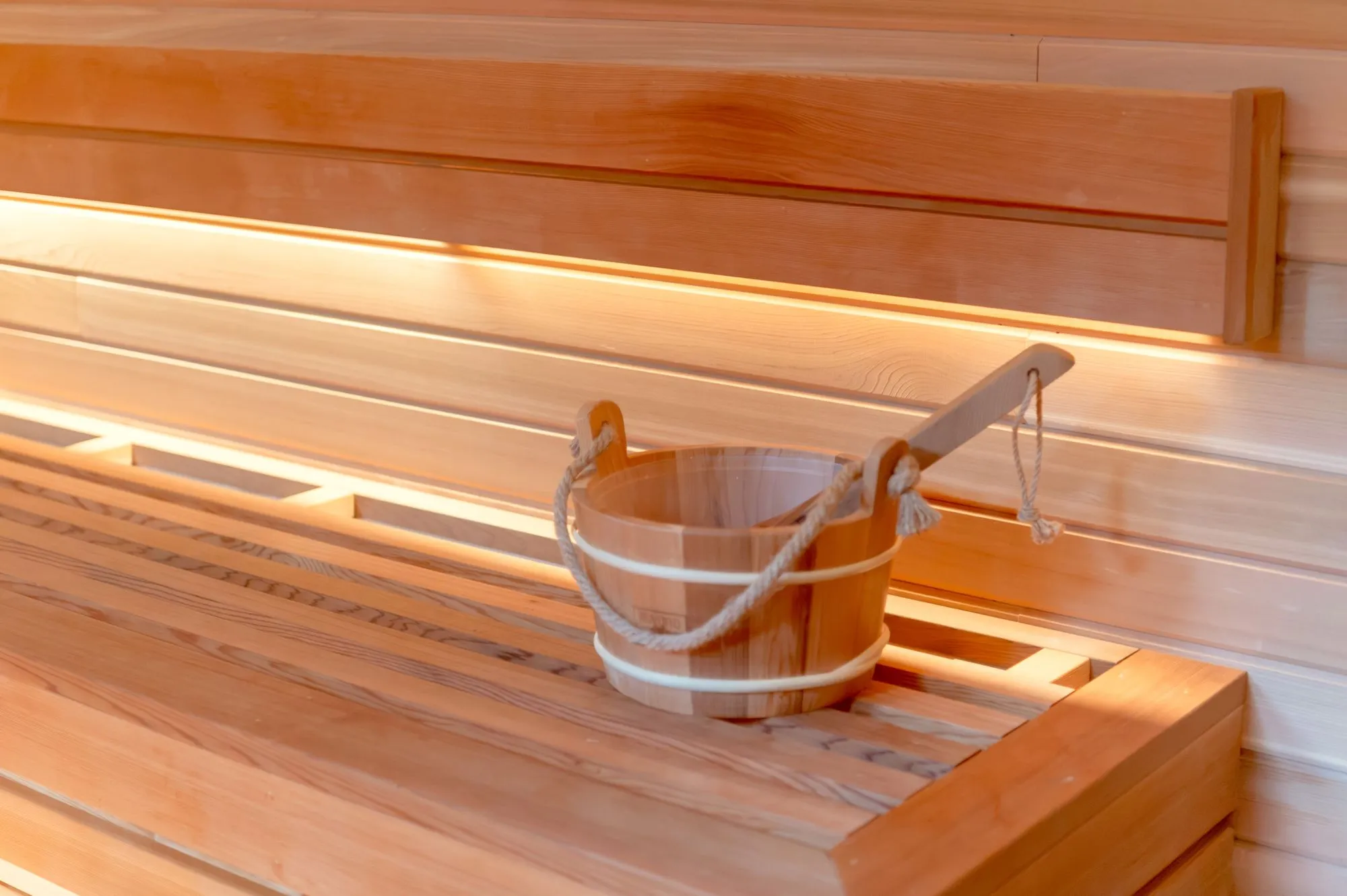 Banquette de sauna en bois clair et seau en bois sous une lumière chaude